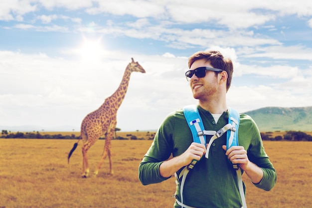 travel, tourism and people concept - happy young man in sunglasses with backpack over giraffe in african savannah background