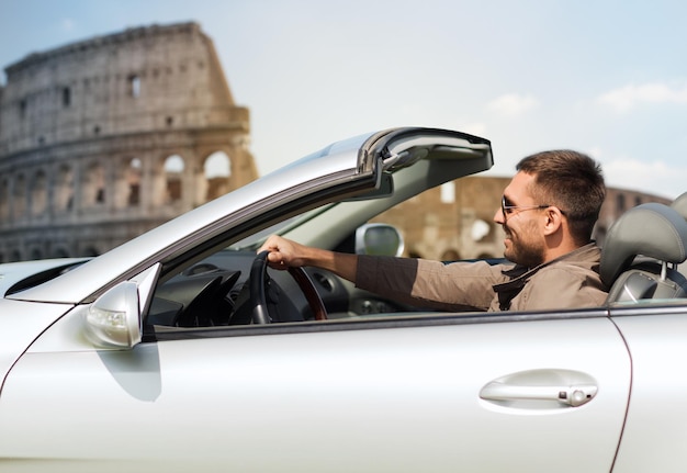 travel, tourism, road trip, transport and people concept - happy man driving cabriolet car over coliseum in rome background