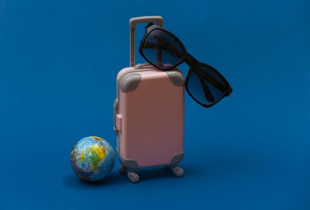 Travel or trip concept. Mini plastic travel suitcase with sunglasses, globe on classic blue background.