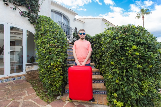 Travel, trip and holidays concept - young man with red suitcase in sunny glasses standing on stairs