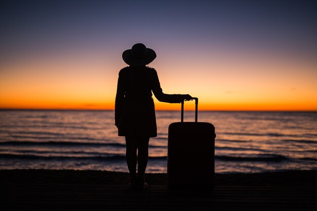 Travel, vacation and summer trip concept - silhouette of young woman in summer dress and hat looking to a sea.