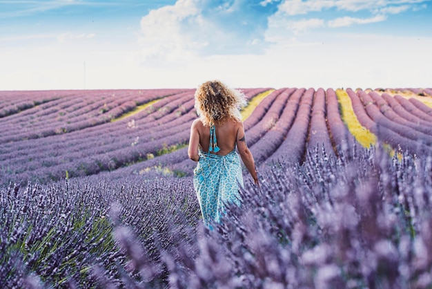 Travel wanderlust lifestyle people concept Back view portrait of woman walking in lavender field wearing blue trendy boho dress Blue sky in background Concept of vacation in scenic destination