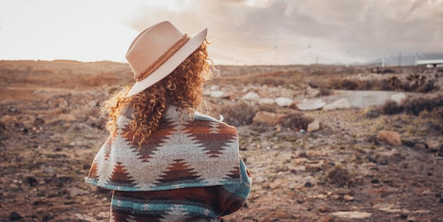 砂漠の風景に沈む夕日を眺めるトレンディな帽子とジャケットを持つ女性の後ろ姿で旅行女性のコンセプトライフスタイル旅行者の人々とアウトドアレジャー活動のコンセプト