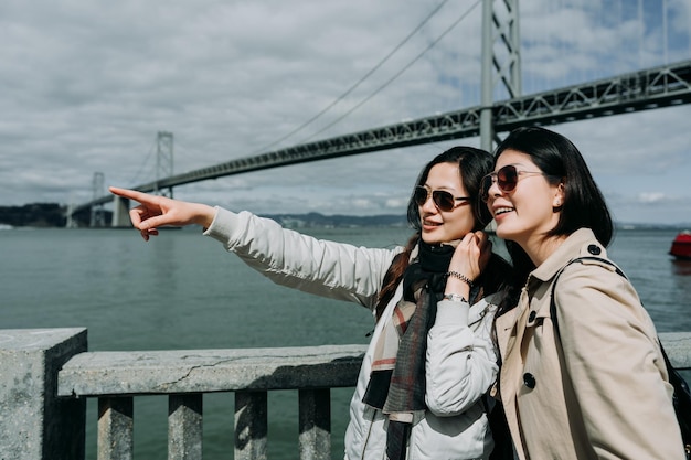 Travel woman pointing with finger outdoors and showing\
something to friend. young backpacker asian sisters tourism in san\
francisco oakland bay bridge on spring holidays. girls happy seeing\
nature