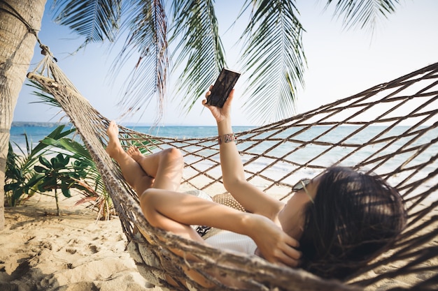 La donna asiatica del viaggiatore che per mezzo del telefono cellulare e si rilassa in amaca sulla spiaggia in koh mak thailandia