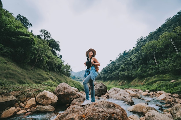 Donna asiatica del viaggiatore con lo zaino che si rilassa e che gode della vista del fiume della montagna