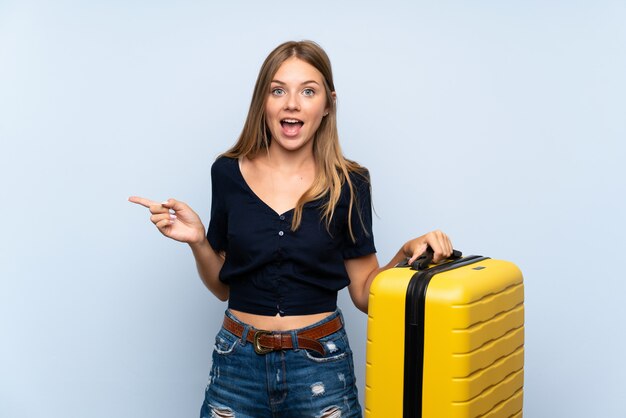 Traveler blonde woman with suitcase surprised and pointing finger to the side