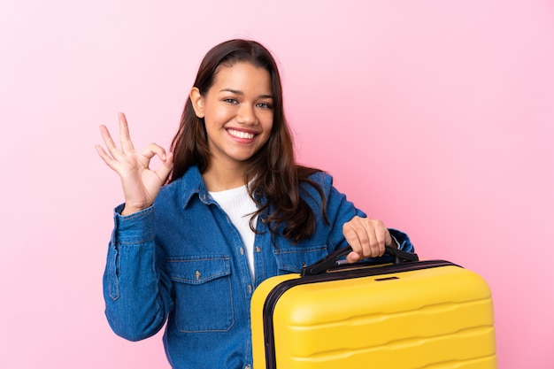 Traveler Colombian woman holding a suitcase over isolated pink wall showing ok sign with fingers