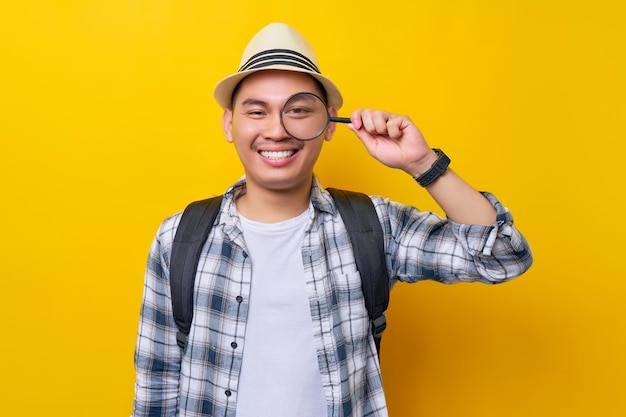 Traveler fun explorer tourist Asian man in casual clothes hat looking through magnifier loupe isolated on yellow background Air flight journey concept