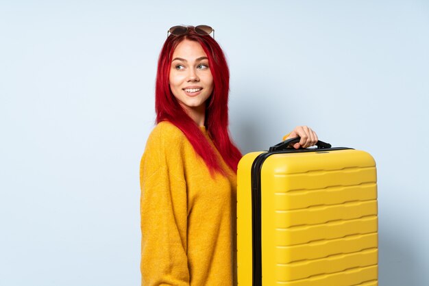 Traveler girl holding a suitcase on blue wall with arms crossed and happy