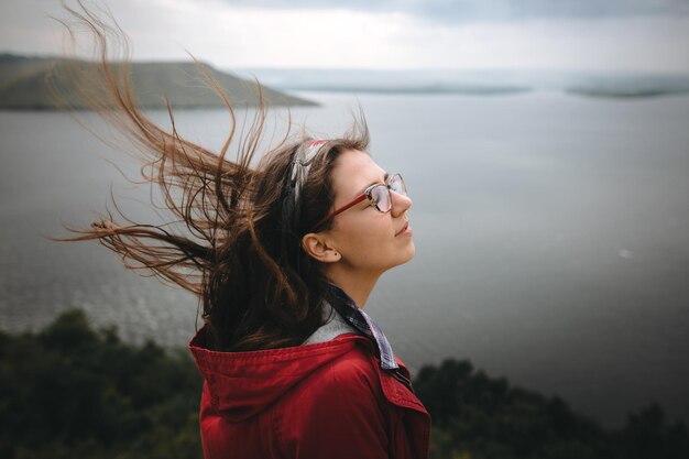 Traveler girl in red raincoat and with windy hair standing on top of rock mountain enjoying beautiful view on river Young carefree woman in glasses relaxing on cliff Copy space Explore