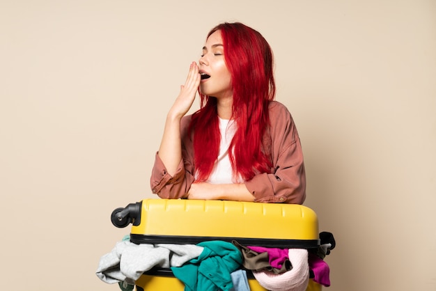 Traveler girl with a suitcase full of clothes on beige wall yawning and covering wide open mouth with hand