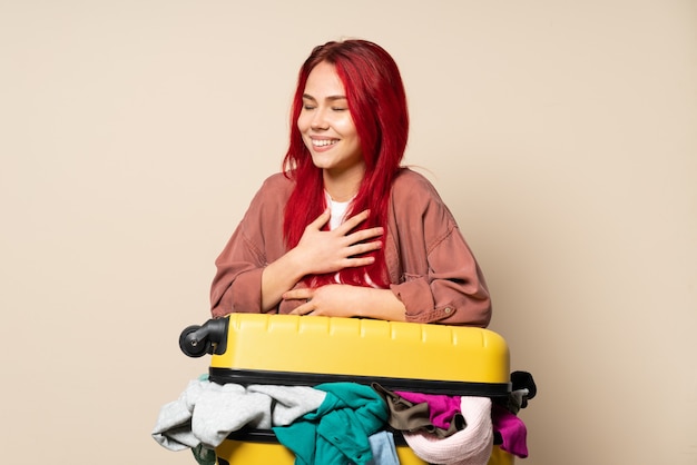 Traveler girl with a suitcase full of clothes isolated on beige background smiling a lot