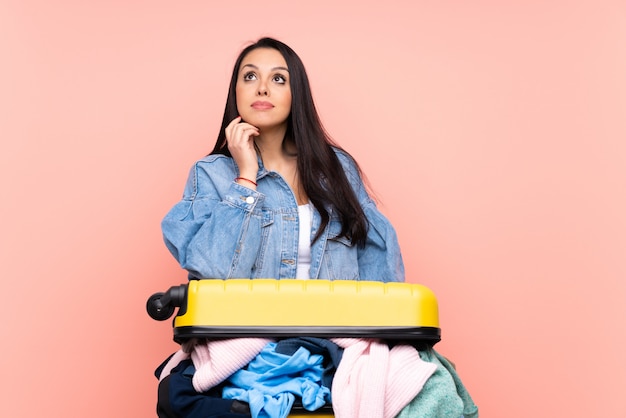 Traveler girl with a suitcase full of clothes over pink thinking an idea