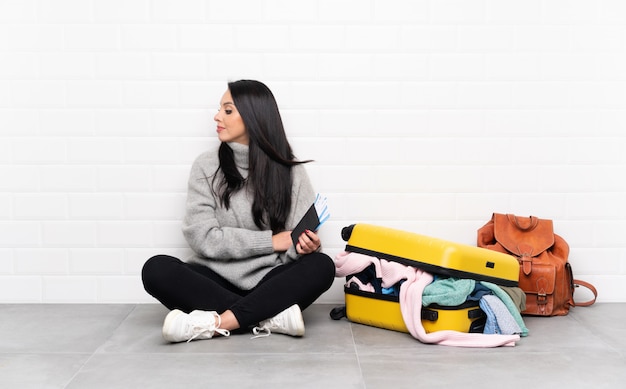 Девушка-путешественница с чемоданом, полным одежды, сидит на полу и смотрит в сторону