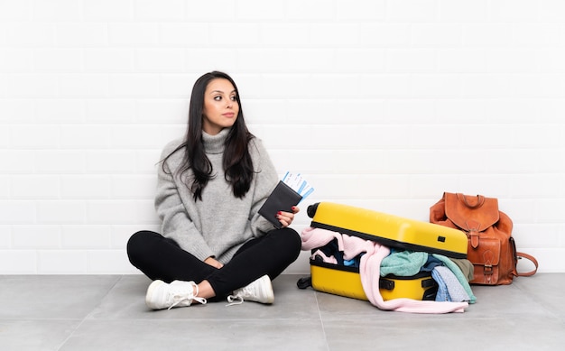 Девушка-путешественница с полным чемоданом одежды сидит на полу, стоит и смотрит в сторону