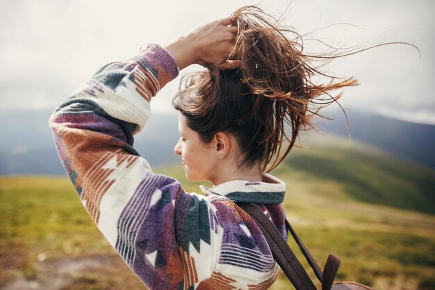 Traveler hipster girl with windy hair and backpack standing on top of sunny mountains space for text stylish woman waving hair atmospheric moment travel and wanderlust concept