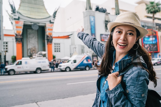 путешественница радостно поднимает руки и показывает на камеру Китайский театр. красивая женщина в синей куртке. самостоятельное путешествие с посещением Голливуда.