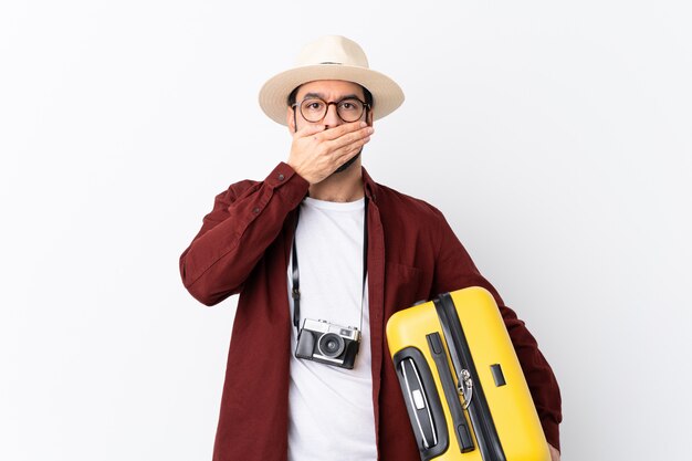 Traveler man man with beard holding a suitcase over isolated white wall covering mouth with hands