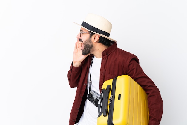 Traveler man man with beard holding a suitcase over isolated white wall shouting with mouth wide open to the lateral