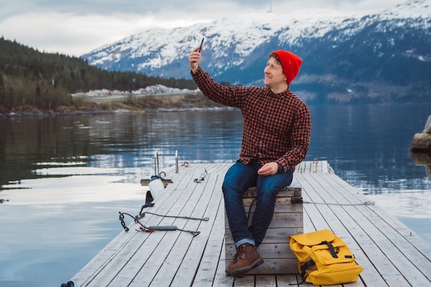 Traveler man taking selfportrait a photo with a smartphone sitting on wooden pier