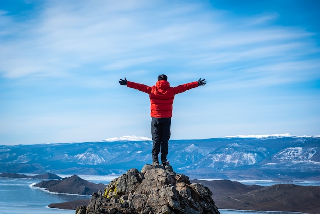旅行者の男は赤い服を着て、日中バイカル湖、シベリア、ロシアで山の上に立って腕を上げる。