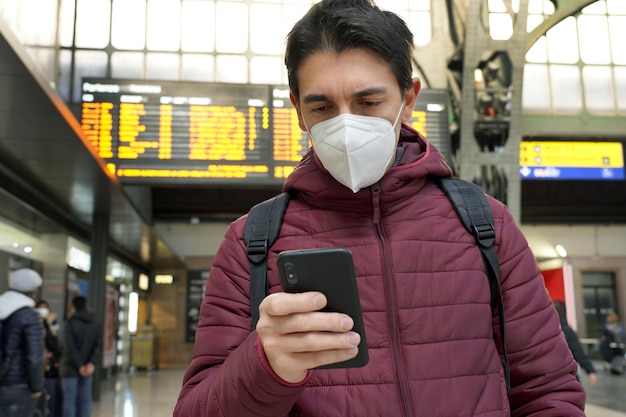 Traveler man wearing kn95 ffp2 protective face mask at the\
airport young caucasian man with behind timetables board of\
departures arrivals checking online ticket of his flight