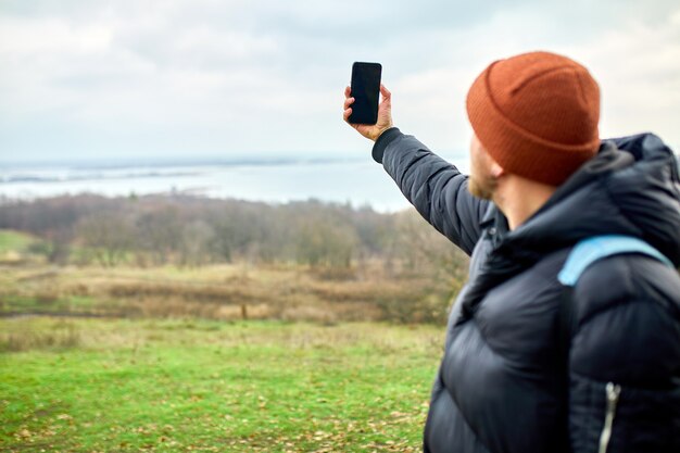 バックパックを持った旅行者の男性は、スマートフォンで写真やセルフィーを作成します。彼の携帯電話は川と山のライフスタイルです。