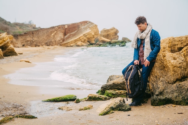 traveler man with a backpack stands on a sandy beach on a background of the sea with rocks