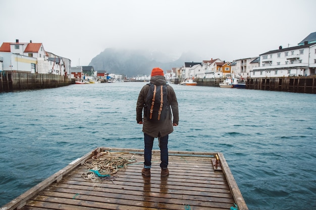 Traveler man with a backpack wearing a red hat standing on the background of fishing houses, ships, mountain and lake wooden pier. Travel lifestyle concept. Shoot from the back.