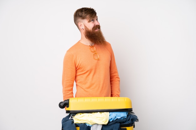 Traveler man with a suitcase full of clothes over isolated white wall laughing and looking up