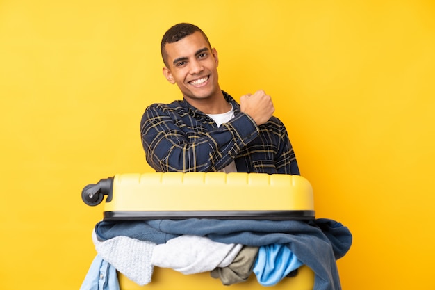 Traveler man with a suitcase full of clothes over isolated yellow wall celebrating a victory