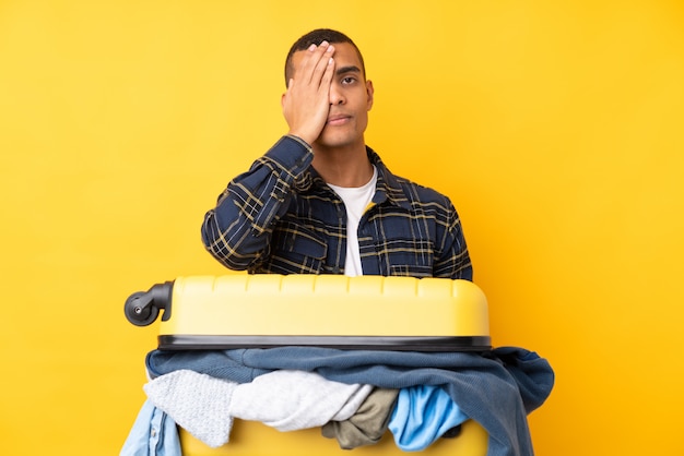 Человек путешественника с чемоданом, полным одежды, над изолированной желтой стеной, закрывающей глаз рукой