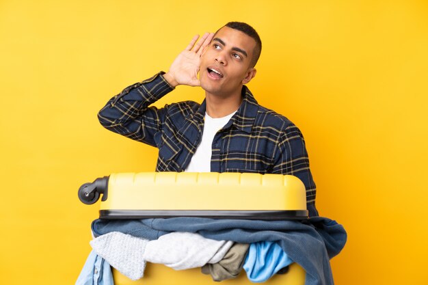 Traveler man with a suitcase full of clothes over isolated yellow wall listening something