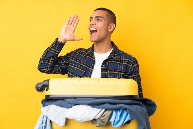 Traveler man with a suitcase full of clothes over isolated yellow wall shouting with mouth wide open