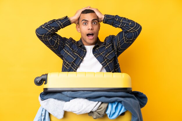 Traveler man with a suitcase full of clothes over isolated yellow wall with surprise facial expression