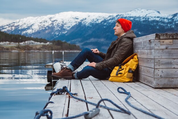 traveler man with a yellow backpack sitting on wooden pier on the background of mountain and lake