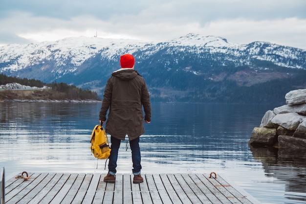 Traveler man with a yellow backpack standing on wooden pier on the background of mountain and lake