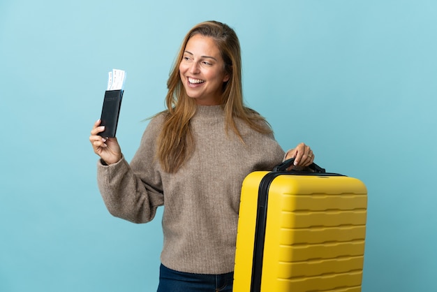 Traveler middle age woman holding a suitcase isolated on blue wall in vacation with suitcase and passport