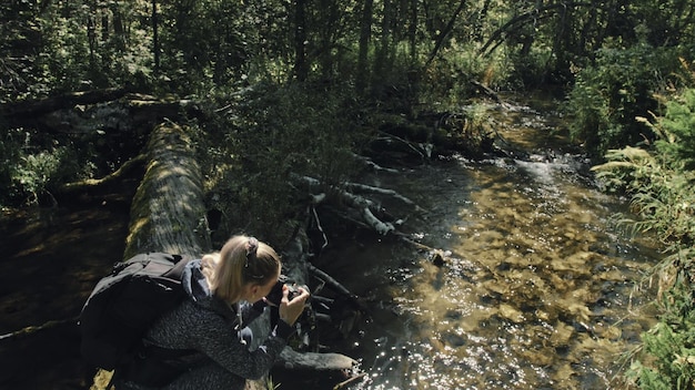 Traveler photographing scenic view in forest river wood bridge\
fallen tree one caucasian woman shooting nice magic look girl take\
photo video on camera photographer travel with backpack\
outdoor