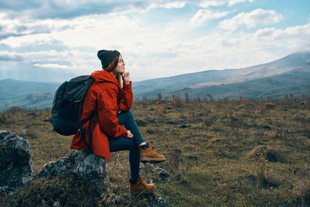 バックパックと赤いジャケットの帽子をかぶった旅行者は、自然の山の石の上に座っています