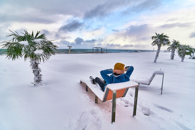 Traveler relaxing on a deck chair among evergreen tropical palm trees covered with snow