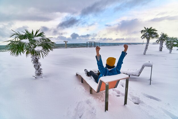 Traveler relaxing on a deck chair among evergreen tropical palm trees covered with snow