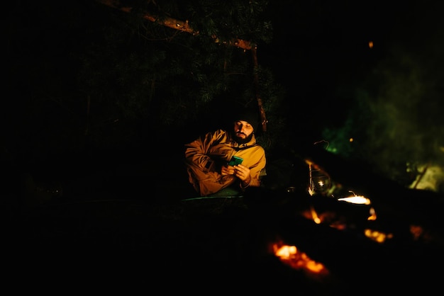 A traveler rests by a night campfire with a phone in his hands A man checks his phone news messages