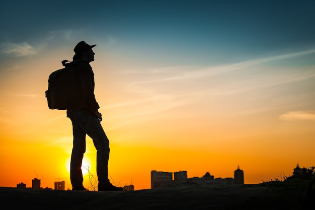 Siluetta del viaggiatore che guarda il tramonto incredibile. giovane uomo casual con zaino e cappello da cowboy in piedi da solo sulla collina sopra il paesaggio urbano serale. stile di vita concetto di viaggio all'aperto sfondo. kiev, ucraina