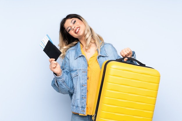 Traveler teenager holding a suitcase isolated on blue wall in vacation with suitcase and passport