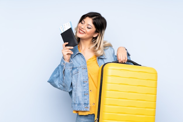 Traveler teenager holding a suitcase isolated on blue wall in vacation with suitcase and passport