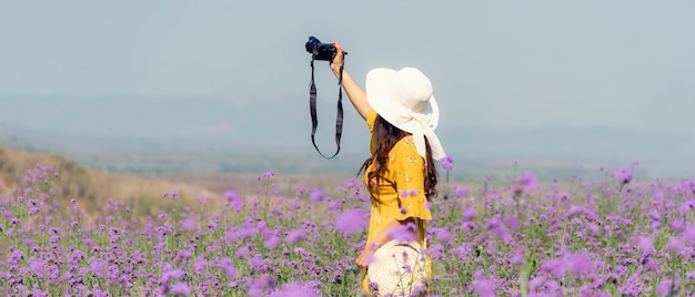 旅行者や観光アジアの女性が立ってカメラを持って紫色のバーベナフィールドで写真の花を撮ります