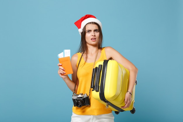Женщина-путешественница в шляпе Санта-Клауса, желтая повседневная одежда держит камеру с сумкой для билетов на синем