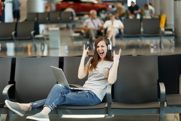 Donna turistica viaggiatrice con cuffie che ascolta musica lavorando su laptop che mostra il segno rock-nroll aspetta nella hall dell'aeroporto internazionale international